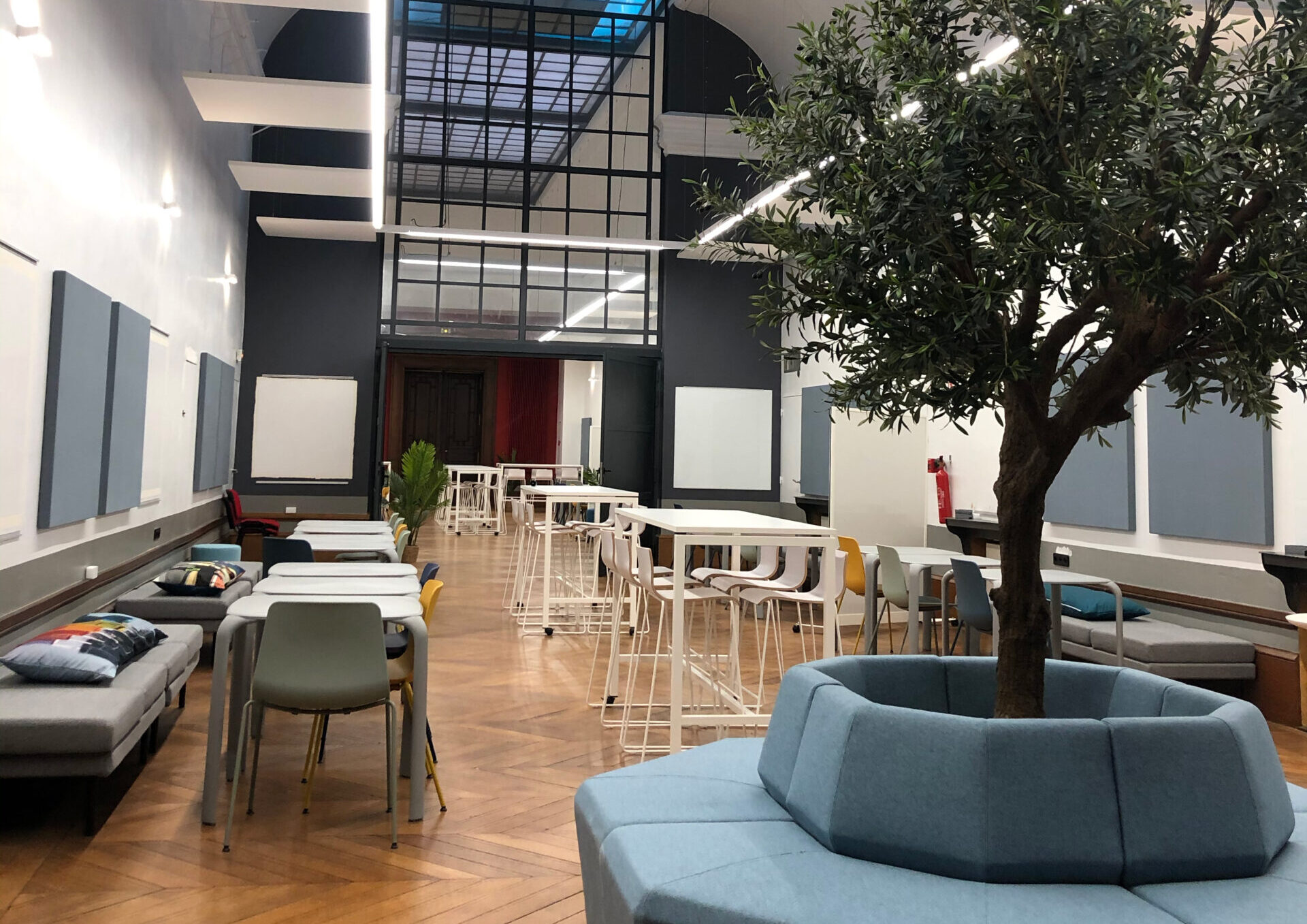 Salle de coworking vide de l’ENSAIT avec haut plafond, tables et chaises, espaces de repos et olivier au premier plan.